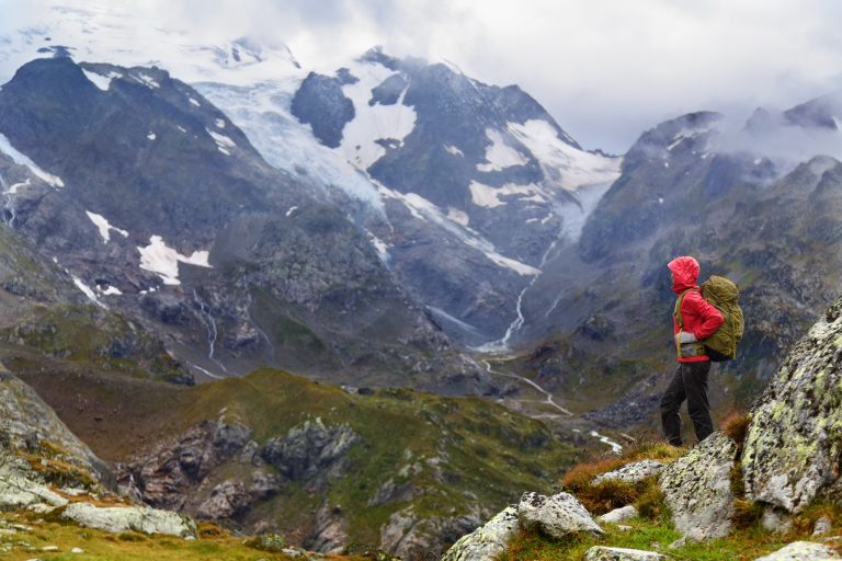 Hiker in a hooded waterproof shell, hiking pants and a loaded pack, looking out over a glacier valley in the rain