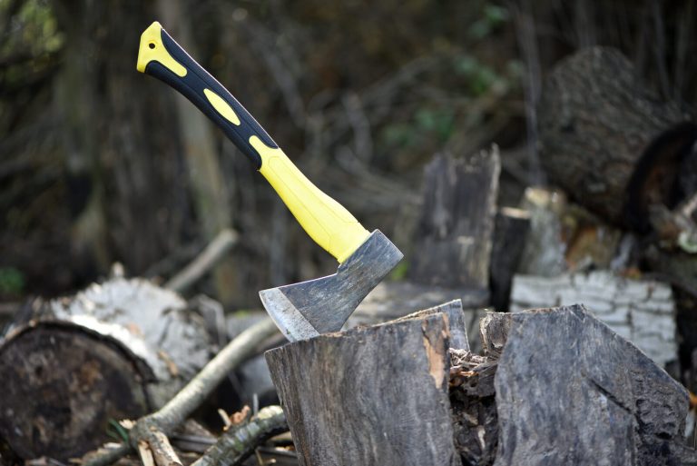 A hatchet with a yellow handle standing embedded in a wooden log outdoors, ready for splitting firewood