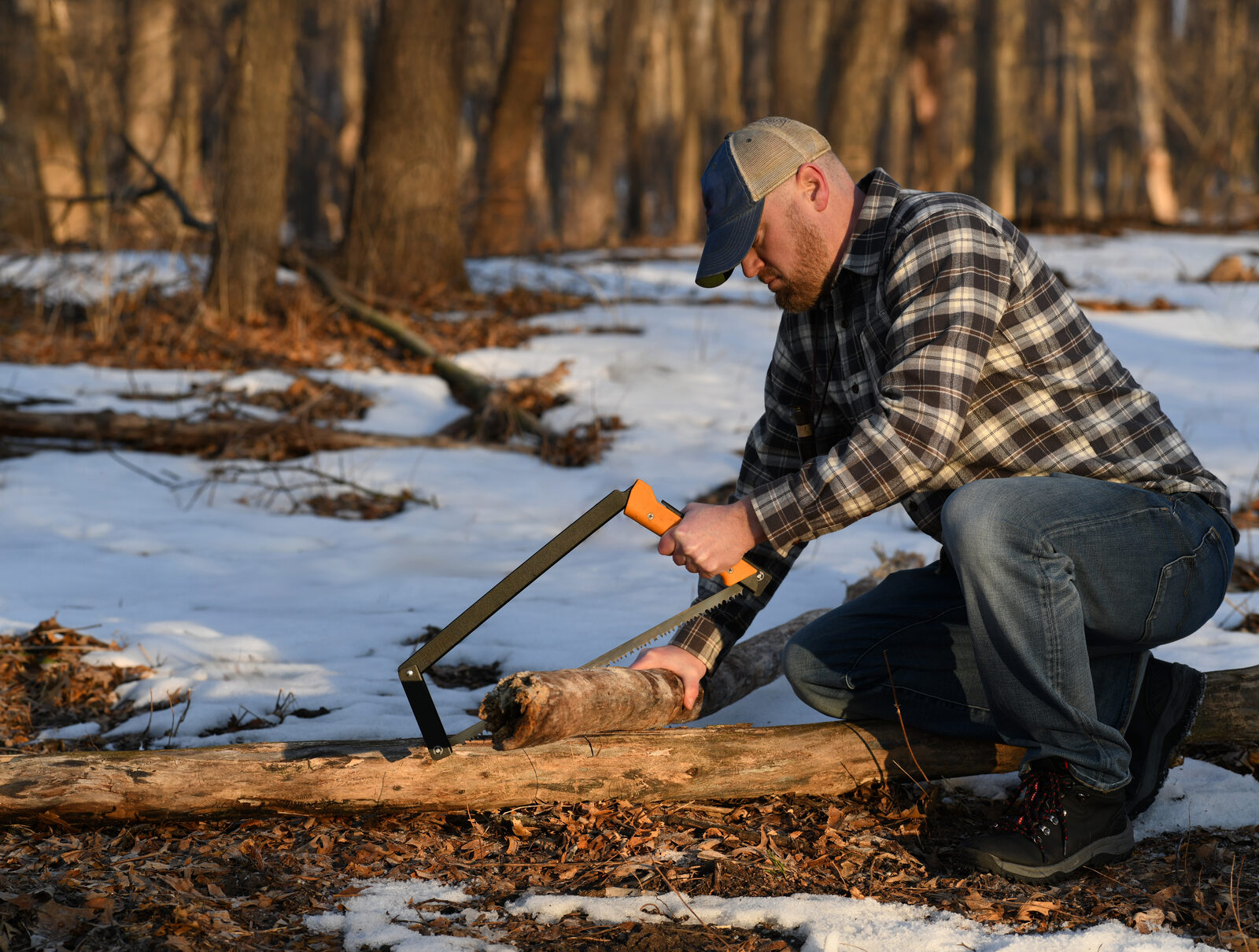 A man in a flannel shirt bucking a log with a bow saw in a snowy forest