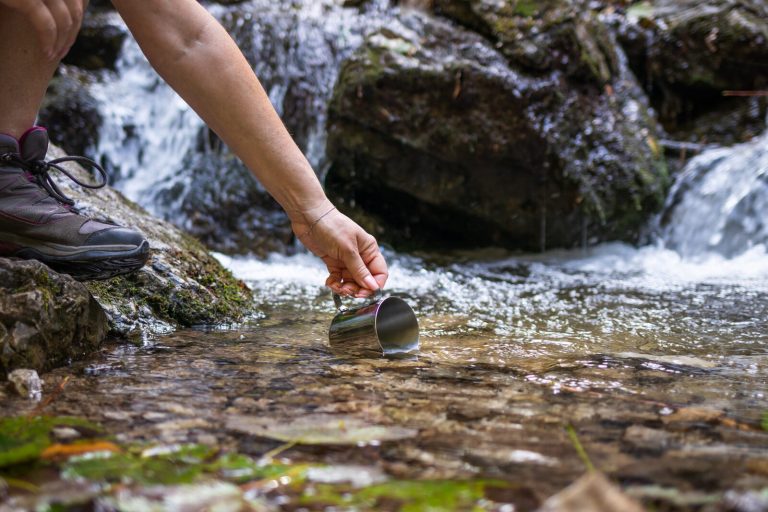 A hiker dips a metal cup into a clear stream below a small waterfall to collect drinking water