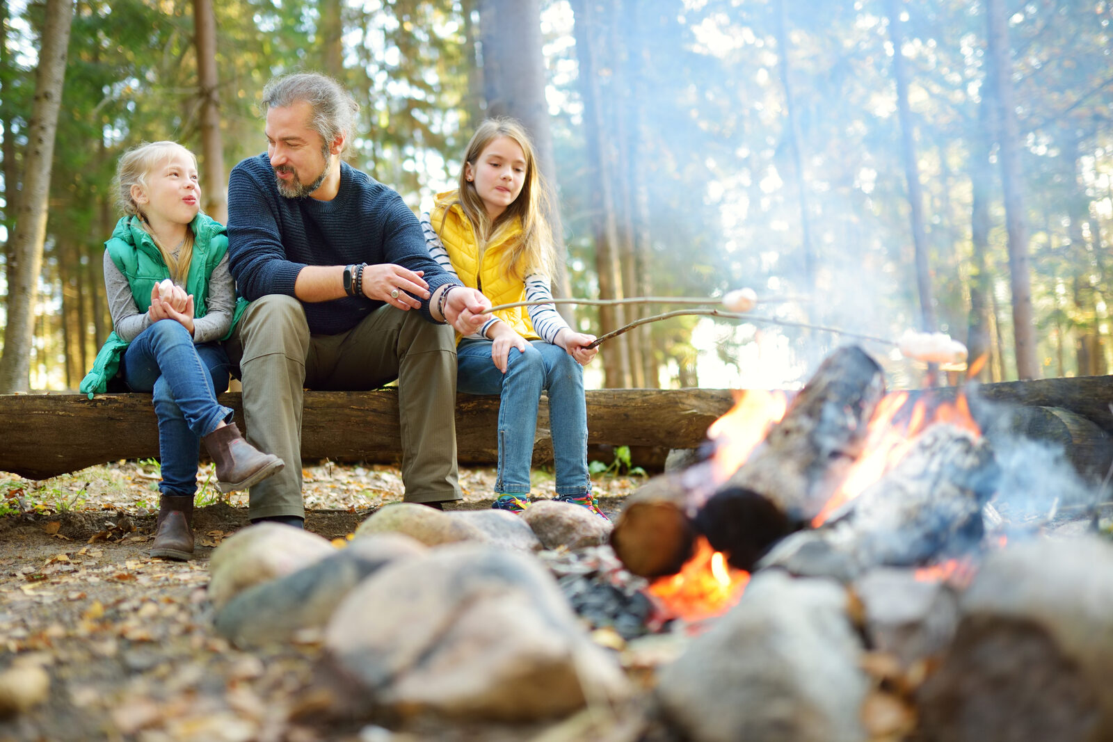 Two children and their father roasting marshmallows on sticks over a campfire in a fall forest