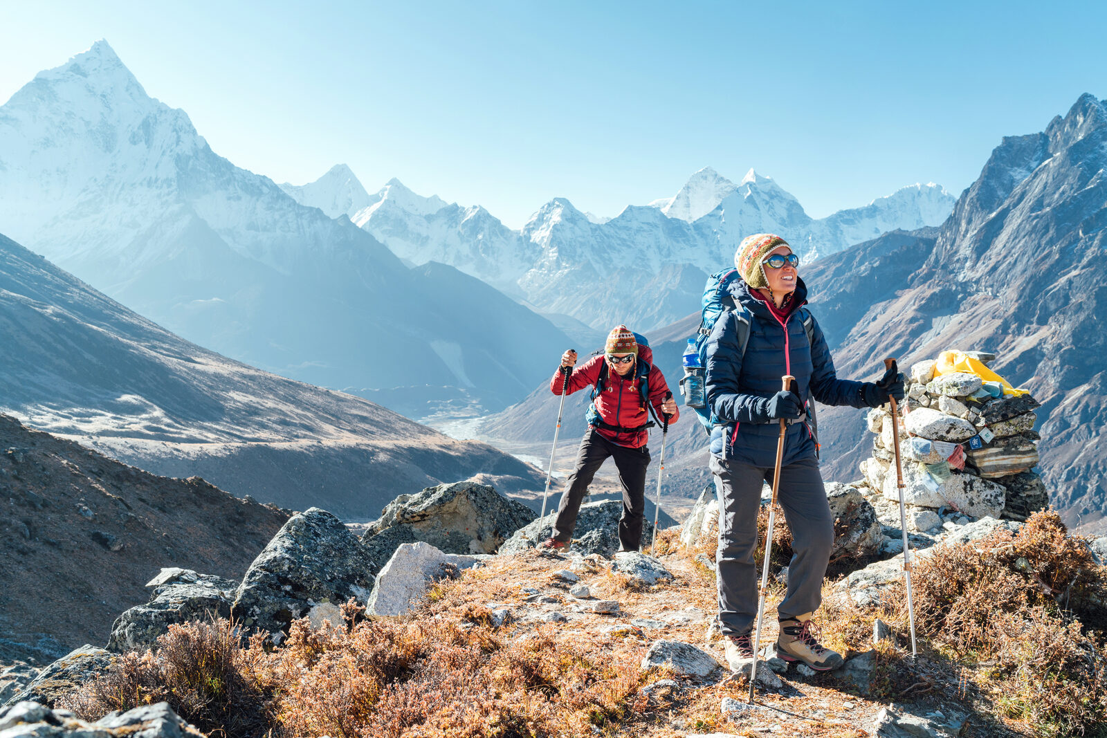 Two backpackers using trekking poles on a rocky ridge with snow-capped Himalayan peaks behind