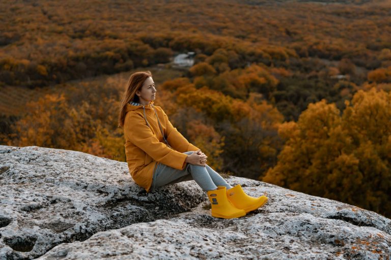 Woman hiker in a waterproof jacket and base layer sitting on a rocky overlook above an autumn forest