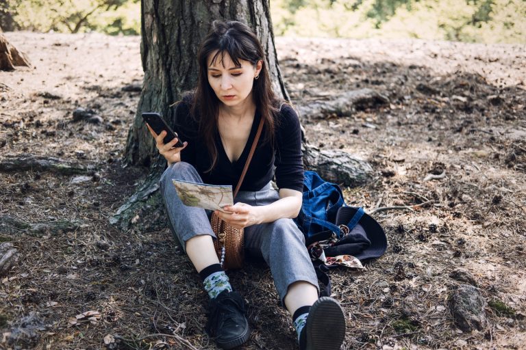 Hiker sitting against a tree in forest, phone in one hand and an open paper topographic map on her lap