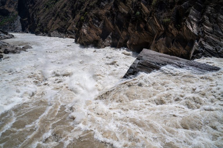 A swollen river tearing through a dark rocky gorge, churning pale water piling against an angular rock slab with calmer water in its lee