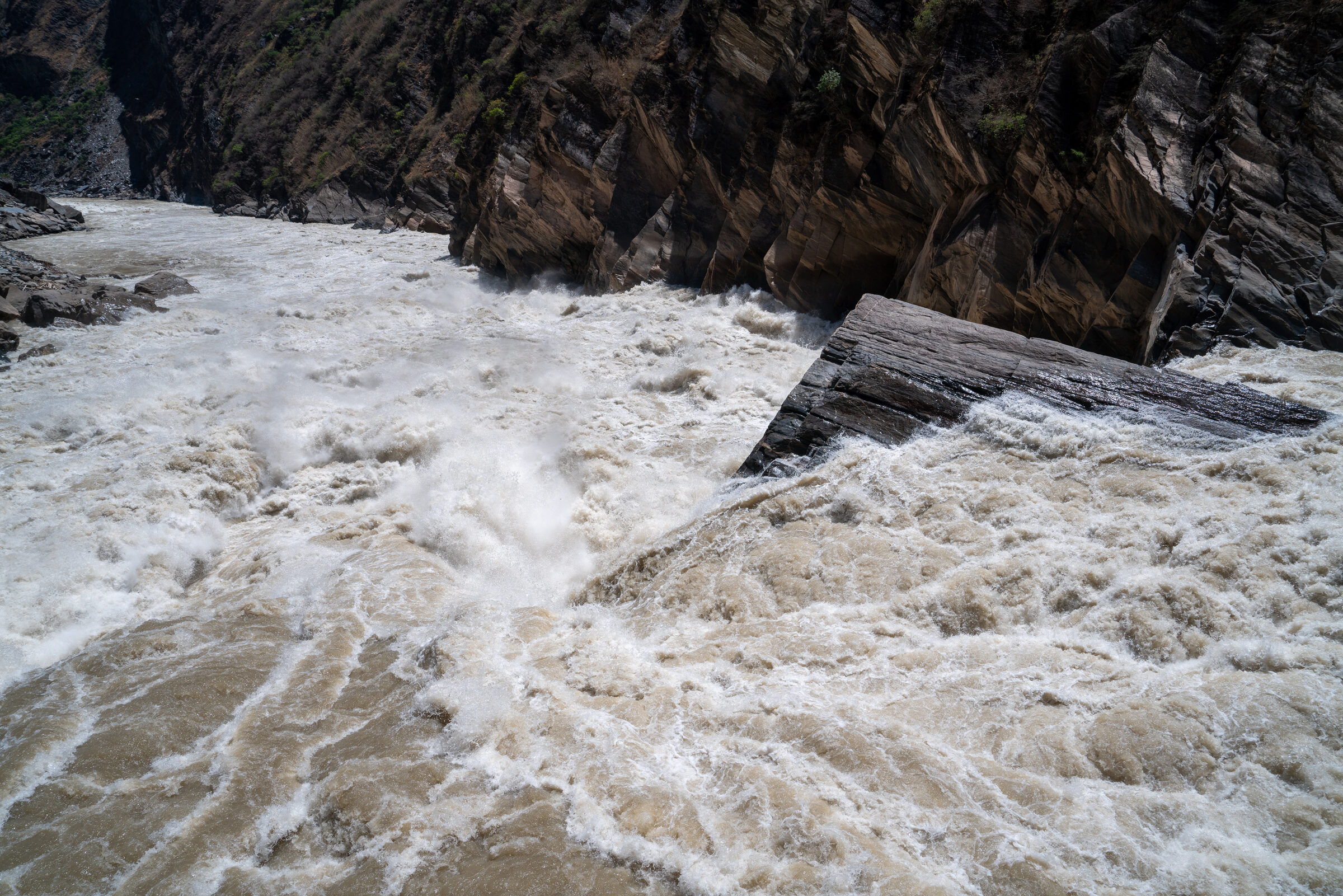 A swollen river tearing through a dark rocky gorge, churning pale water piling against an angular rock slab with calmer water in its lee