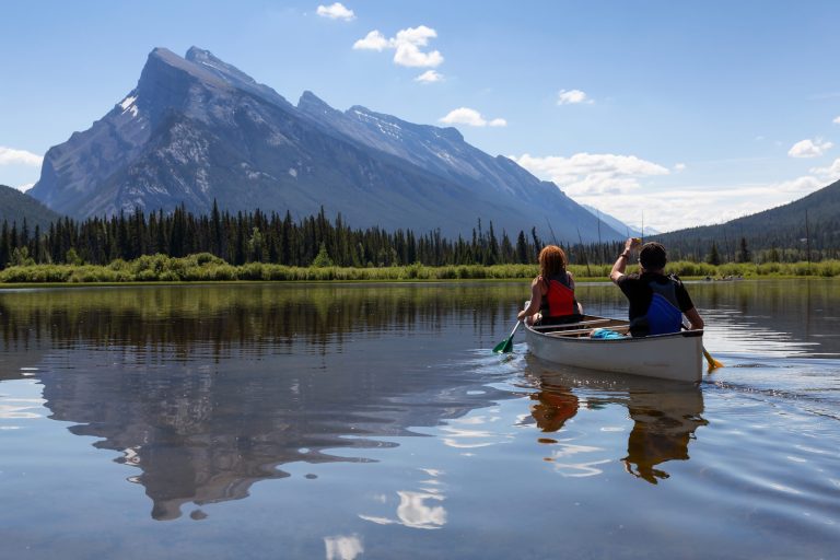 Two paddlers wearing life jackets paddle a tandem canoe across a calm lake below Canadian Rockies peaks