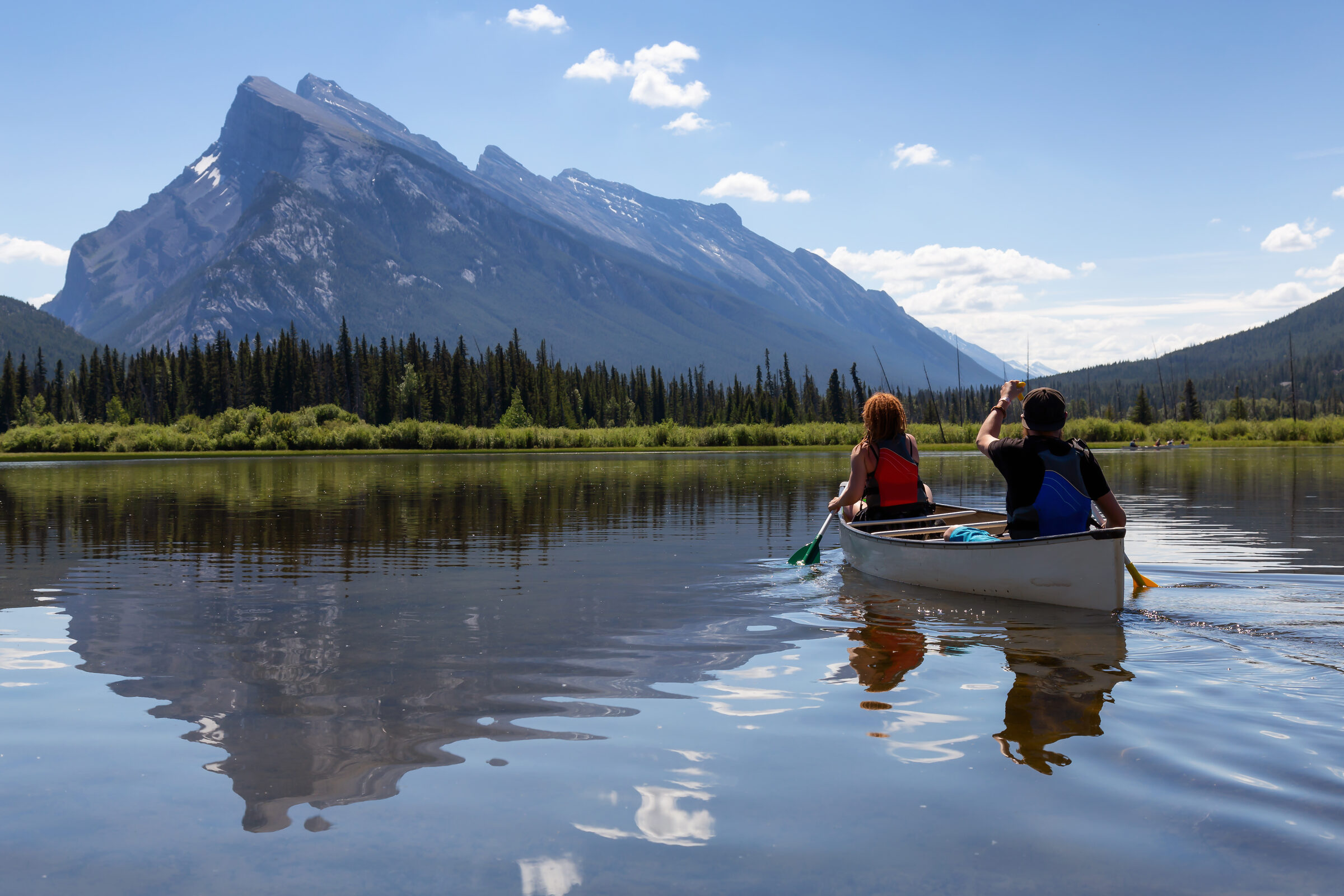 Two paddlers wearing life jackets paddle a tandem canoe across a calm lake below Canadian Rockies peaks