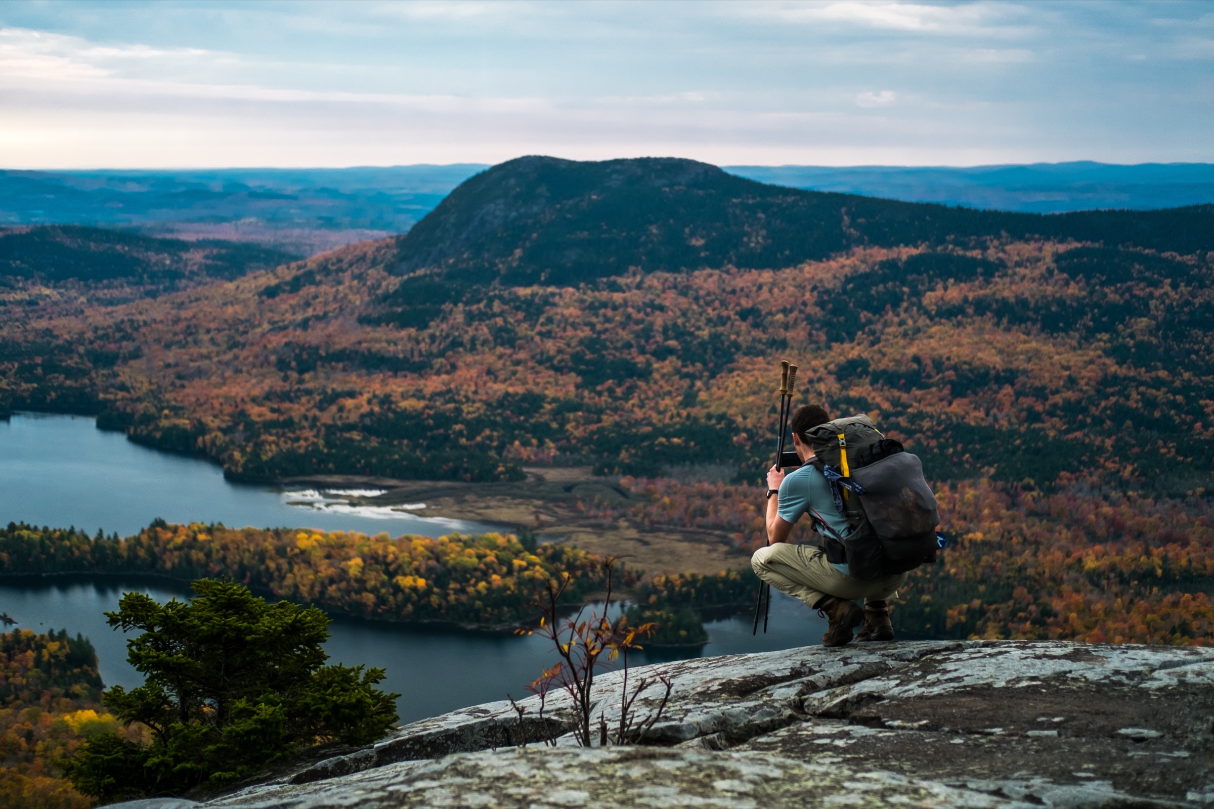 A backpacker with a full multi-day pack and trekking poles crouched on a granite summit ledge, looking out over a lake and autumn forest in Maine on the Appalachian Trail