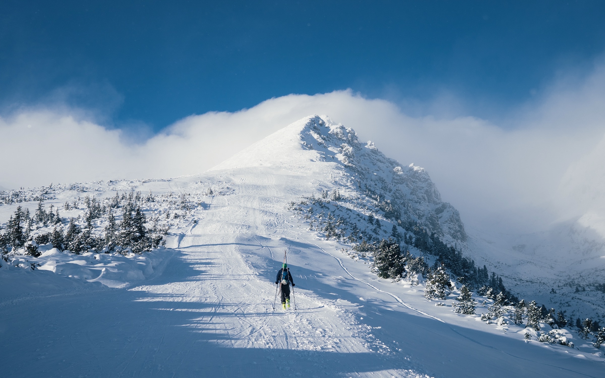 A lone ski tourer walking uphill with skis on their pack toward a steep, snow-covered summit with cloud streaming off the ridge