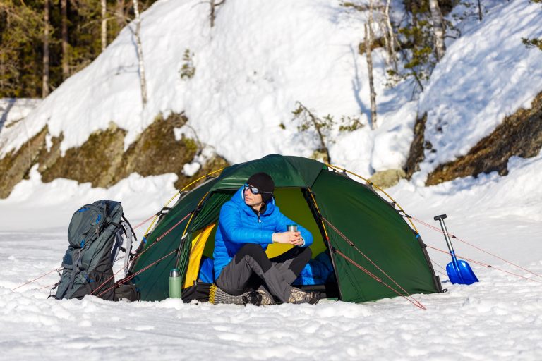 A winter camper in a down jacket sits in the doorway of a dome tent pitched on snow, holding a hot drink, with a large backpack, an insulated bottle, a foam sleeping pad and a snow shovel beside the tent