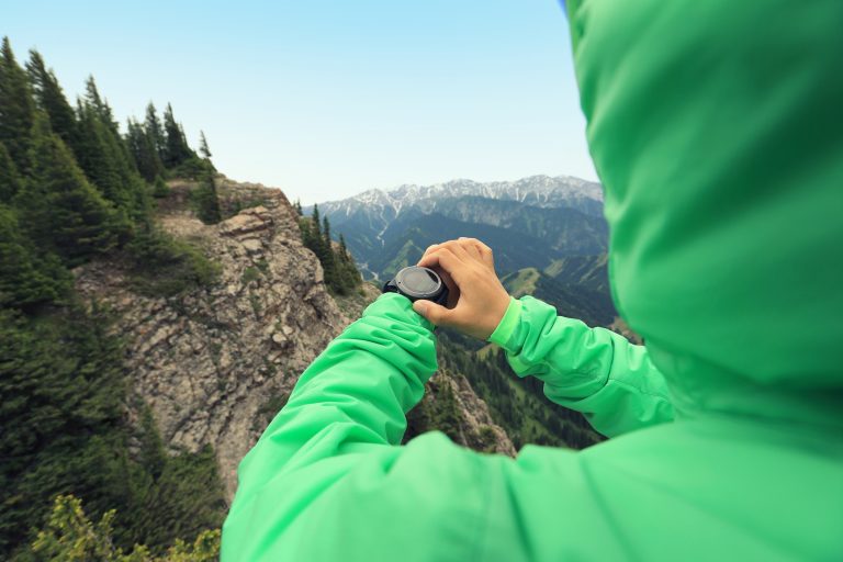 A hiker in a green hooded rain shell checks the watch on her wrist from a rocky viewpoint above a forested mountain valley