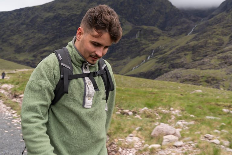 A hiker in a plain green fleece pullover and a daypack with the chest strap fastened, walking in a Scottish mountain glen