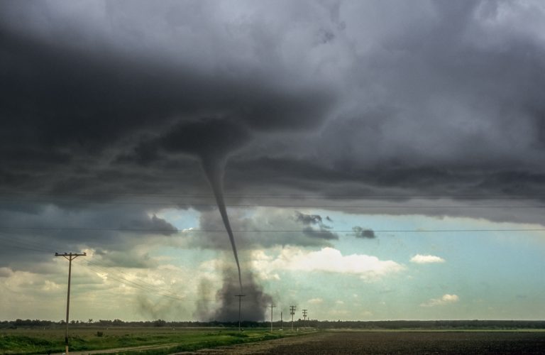 A narrow tornado reaching down from a dark storm cloud to a column of dust over flat farmland, with power poles along a road in the foreground