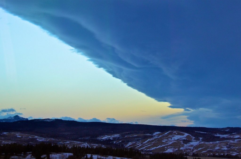 A flat sheet of dark cloud ending in a sharp diagonal edge, with a strip of clear pale sky between the cloud and snow-patched foothills