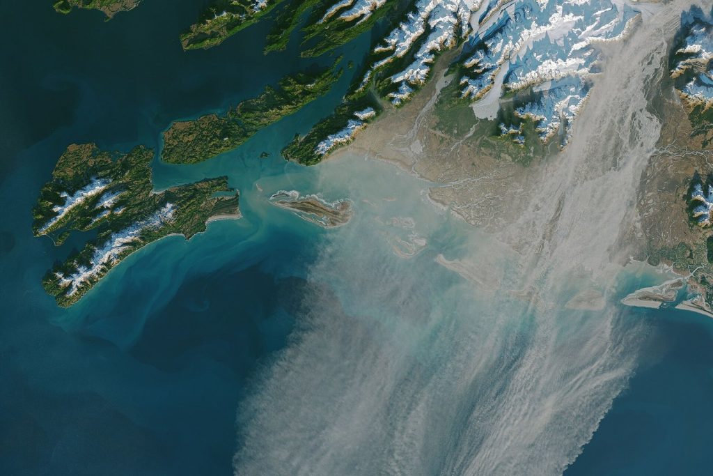 Satellite view of the Alaska coast with a broad grey plume of dust streaming from a river delta between snow-covered mountains out over the sea