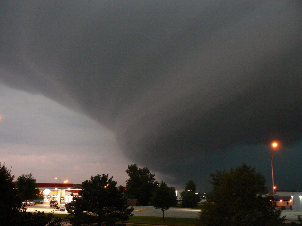 A low, dark, curved shelf of storm cloud filling the sky above trees and a lit petrol station at dusk