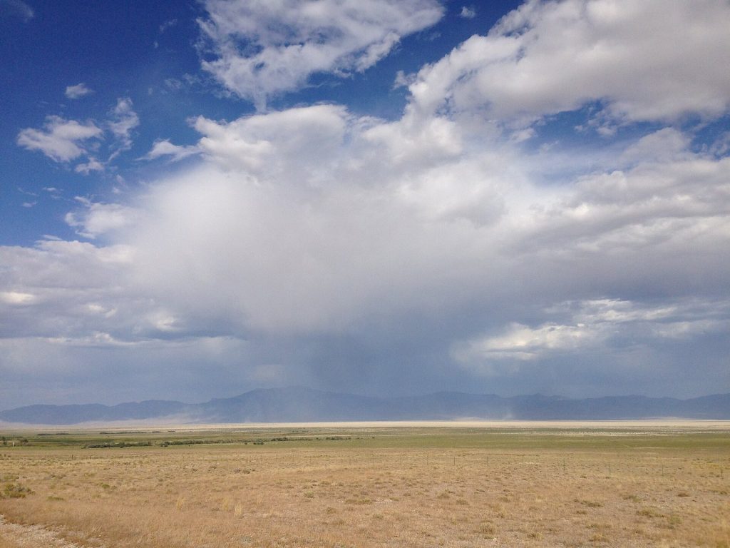 A thunderstorm cloud over a wide dry valley with a pale shaft of rain beneath it that fades before reaching the ground