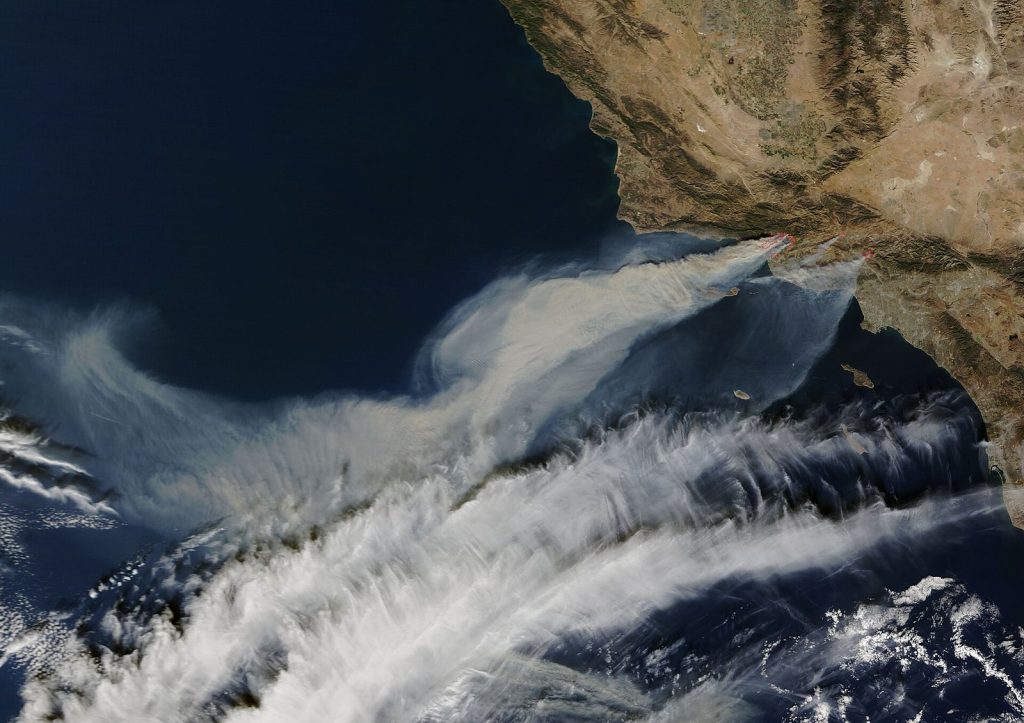 Satellite view of the Southern California coast with long plumes of grey smoke blowing from the land far out over the Pacific Ocean